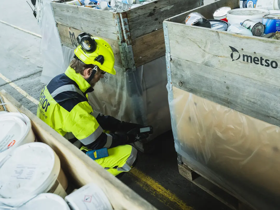 Worker in high-visibility safety gear scans materials in labeled wooden crates containing industrial waste, with "Metso" branding visible on one crate.